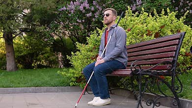 A blind man with a cane sits on a bench in a park