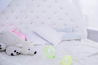 Big white teddy bear on a white big bed with white pillows and multicolored balloons