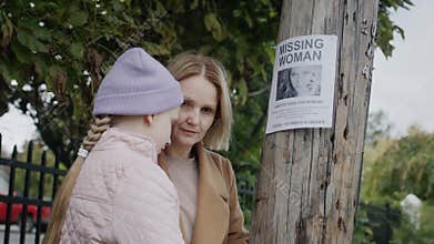 A sad woman with a child stands at a pole where an advertisement for a missing young woman has been announced. Search