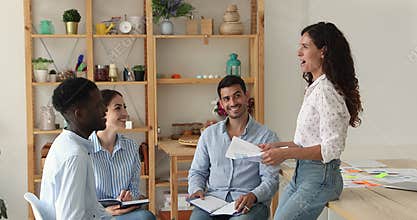 Latin woman sit on desk laugh speak before smiling teammates