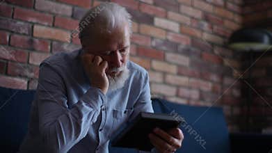 Sad thoughtful grey-haired mature male looking at family portrait holding photo frame sitting on couch at home. Concept