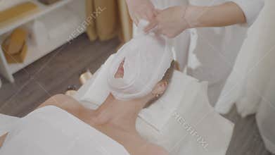 Master spa salon wraps warm towel around client face with massaging movements.