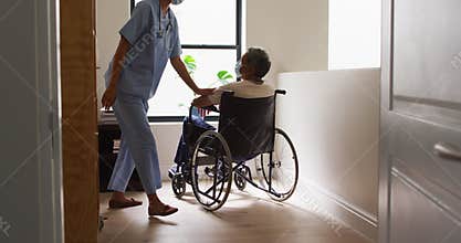 Mixed race female doctor wearing mask assisting senior man in wheelchair at home
