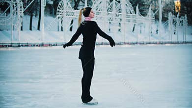 Young blonde woman in black clothes with high ponytail figure skating on the public ice rink in the morning