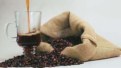 Pour fresh brewed coffee into a clear mug. A burlap bag with coffee beans.