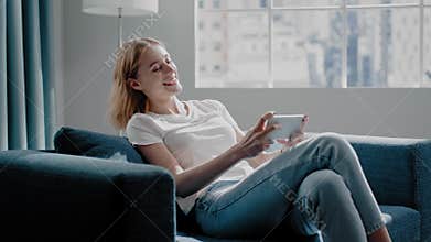 Joyful woman plays game on smartphone sitting on azure sofa