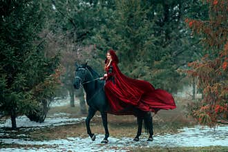 Medieval woman princess in red dress sits astride black steed horse. Girl rider in vintage cloak cape train flies in