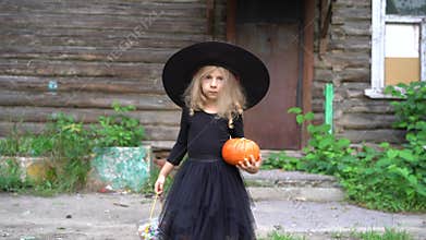 a little girl in a black carnival sorceress costume and hat, with a pumpkin in her hands near the old house, the concept