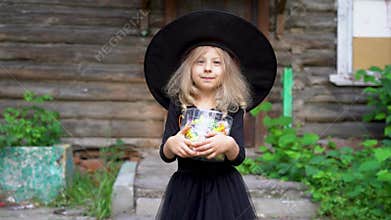 a little blonde girl in a black masquerade costume for halloween with candy against the background of an old house