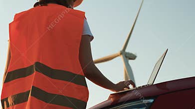 An windmill engineer supervises the work of the wind turbines on the laptop standing near the car. Generation of eco
