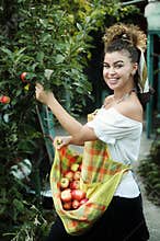 Beautiful woman with curly brown hair holding apples in apron in the garden