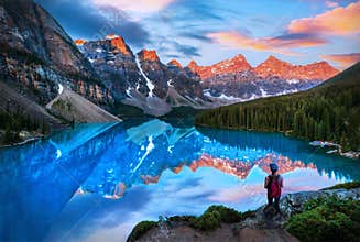 Hiker watching the sun-lit mountains of a Summer sunrise over glacier-fed Moraine Lake in Banff, Canada