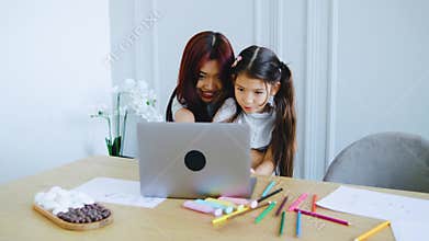 Smiling brightly, a mom and her child wave at a laptop during a video call. Their table, with art supplies and flowers