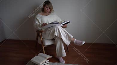 a medium-sized woman in light clothes reads a book while sitting in a white chair in a light room