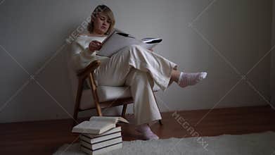 A medium-sized woman in light clothes reads a book while sitting in a white chair in a light room