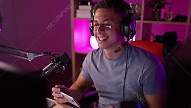 Hispanic man podcasting in a gaming room at night, making a gesture to greet his audience while holding his smartphone
