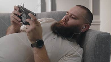 Fat bearded man addicted playing portable console games with happy excitement emotions while lying on couch. Positive