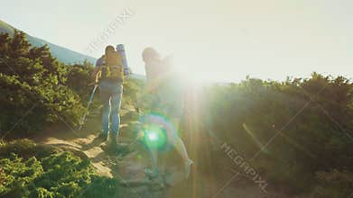 Couple with a backpack with trekking sticks goes up the mountain path. In the rays of the setting sun. Back view