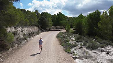 A blond boy runs along a mountain forest road, shooting on a drone from the back.