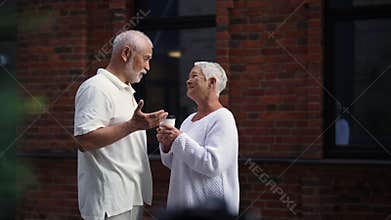 Remote shot of pretty elderly couple enjoying coffee outdoors in city street, talking and smiling happily against brick
