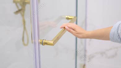 A delicate hand closes a glass shower door with a shiny gold handle, set against a white bathroom backdrop. This photo