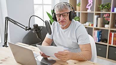 A mature man wearing headphones speaks into a studio microphone, holding a script indoors, suggesting a podcasting or broadcasting