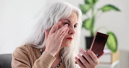 Elderly woman with gray hair looking at wrinkles and bags under eyes in mirror at home 4k movie slow motion