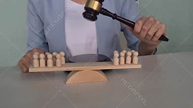 A wooden gavel bangs on a balance scale with wooden people on a white background. The gavel is in the middle