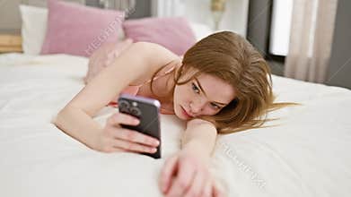 Young woman browsing smartphone while reclining on white bed in a cozy bedroom