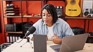 A hispanic middle-aged woman engages in podcasting inside a studio using a microphone, laptop, and tablet