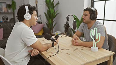 Two men podcasting indoors at a wooden table with microphones and neon cactus light on stage