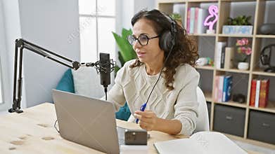 Hispanic woman podcasting in radio studio with microphone and laptop on wooden table