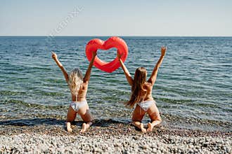 Two women are posing on a beach holding a heart shaped inflatable raft