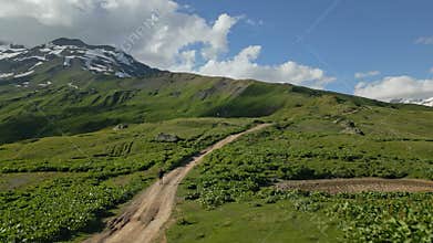 Lone traveler walks along dirt path in alpine meadows of Svaneti mountains. Aerial view of green landscape with snow