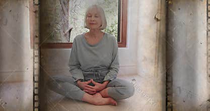 Meditating senior woman in grey loungewear sitting legs crossed at home by window with film borders