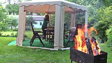 Woman prepare supper table in outdoor bower. Firewood burn