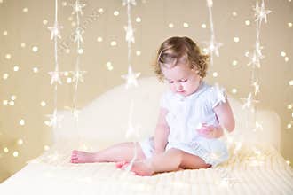 Cute toddler girl with her toy bear on a white bed between beautiful warm Christmas lights