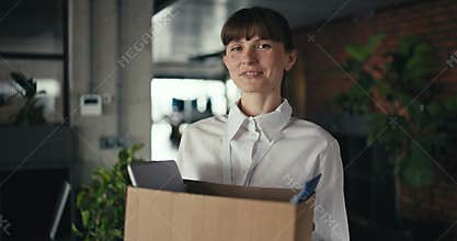 Young professional woman with bangs wearing a white shirt holds a moving box with office supplies during onboarding in a