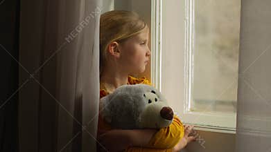 Upset sad little Caucasian girl child kid orphan daughter looking out window hugging teddy bear toy waiting for parent