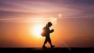 Silhouette of school child walking at sunset with book and backpack.