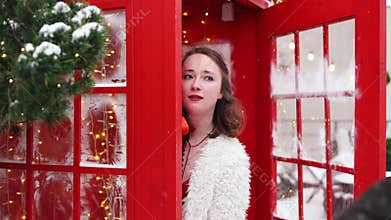 A Christmas concept, Happy woman in a fur coat posing in a red telephone booth on snowing day.