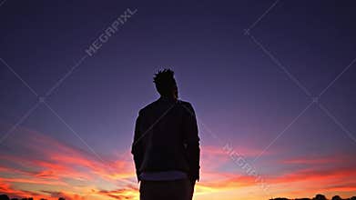 Solitary figure stands beneath a vast sunset sky, embers of dusk bleeding upward into the darkness