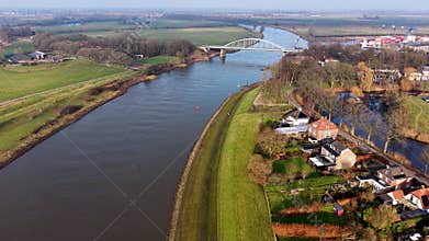 Aerial view of river with cargo ship, arched bridge, and mixed use banks. Residential homes, gardens, and green fields frame a