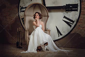 A smiling girl in a wedding dress in strange chair. The bride in a chair on the background of clocks and fireplace tool set.