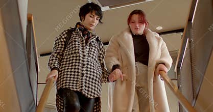 Two women dressed warmly laugh and converse on hotel staircase in cozy lobby