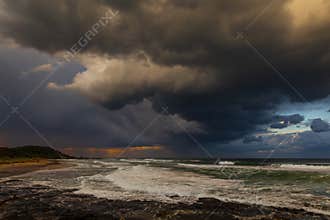 Rain falls from stormy skies over the sea and beach at sunset.