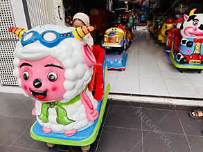 A young child wearing an orange outfit with a beige hijab is seated on a coin-operated ride shaped like a colorful cartoon sheep.