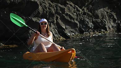 Kayak paddling woman enjoying sea travel along rocky cliff in bright sunshine