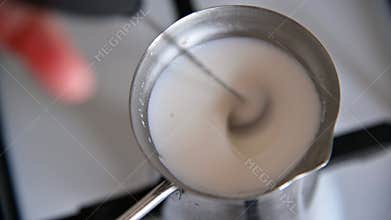 A close-up of milk boiling on a gas stove in a metal coffee pot. A hand-held frother froths the milk. Homemade coffee making.