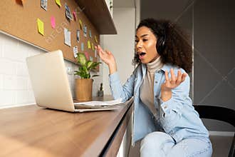 Funny surprised afro american woman wearing headphones video calling on laptop.